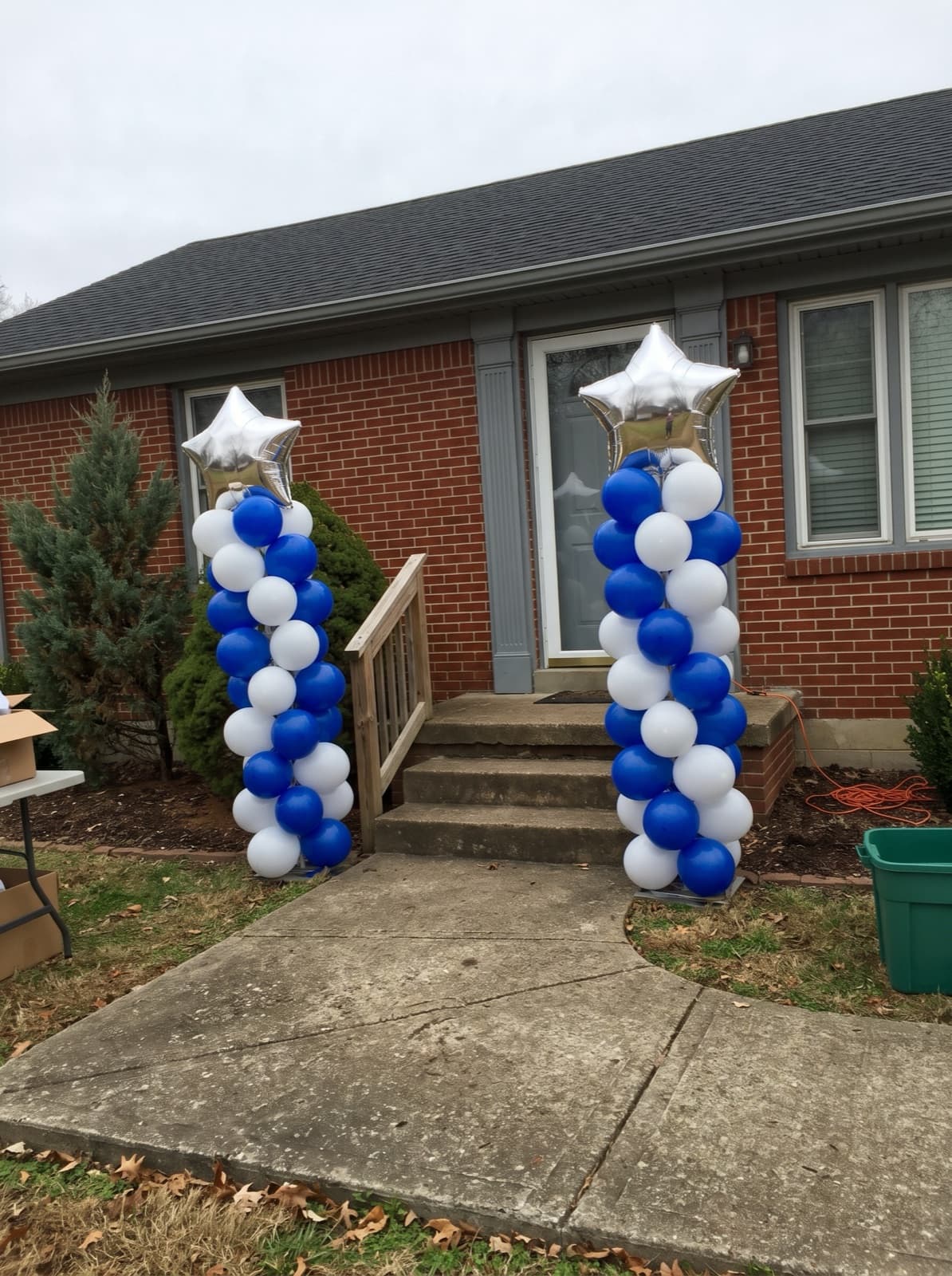 Two royal blue and white spiral balloon columns with silver star toppers beside the front steps of a brick house